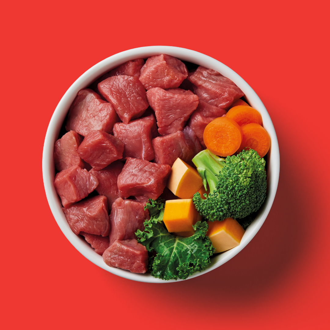 White bowl with raw beef chunks and vegetables on a red background