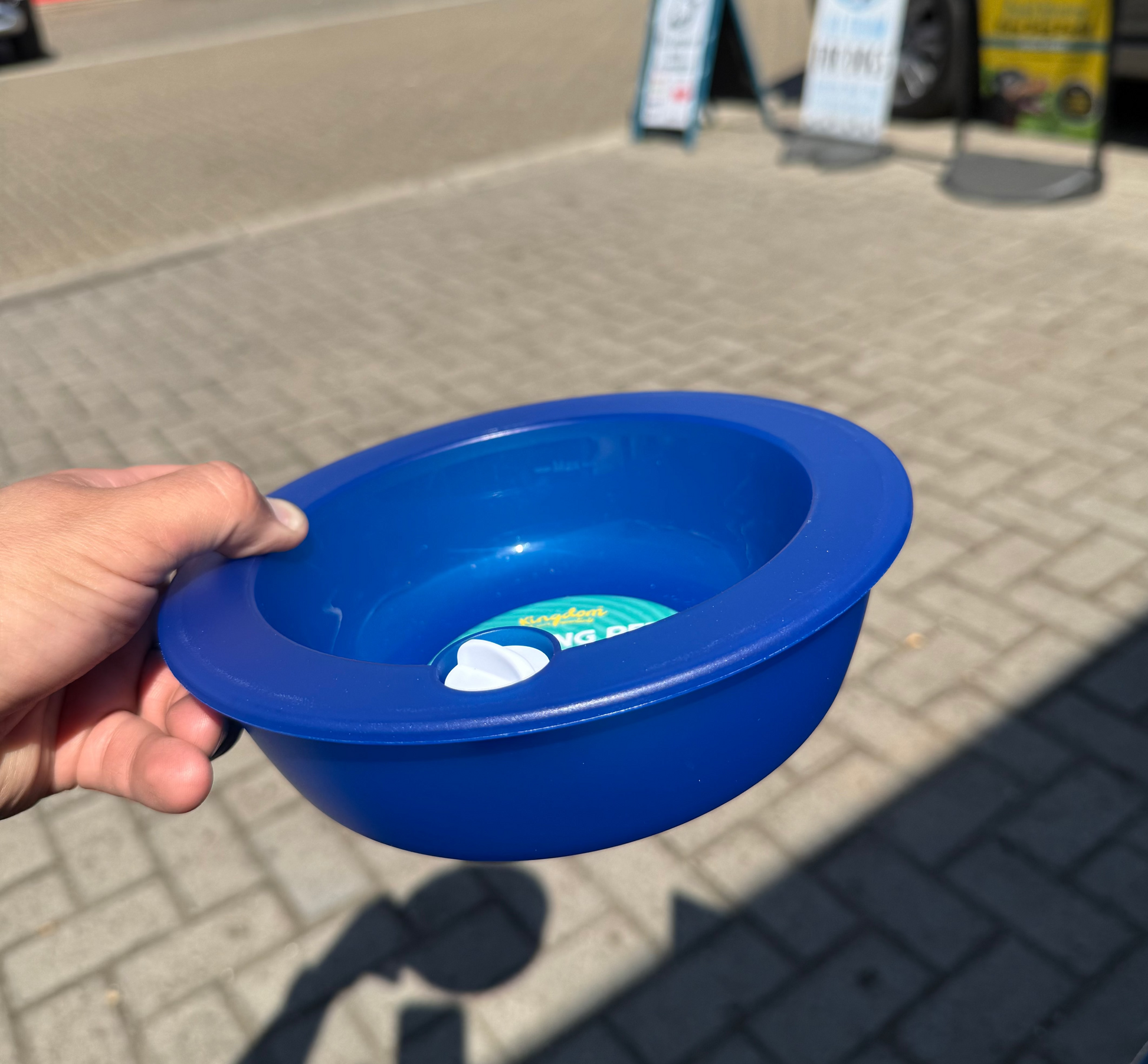 A hand holding a bright blue plastic bowl outdoors on a paved street, with cars and storefronts blurred in the background.