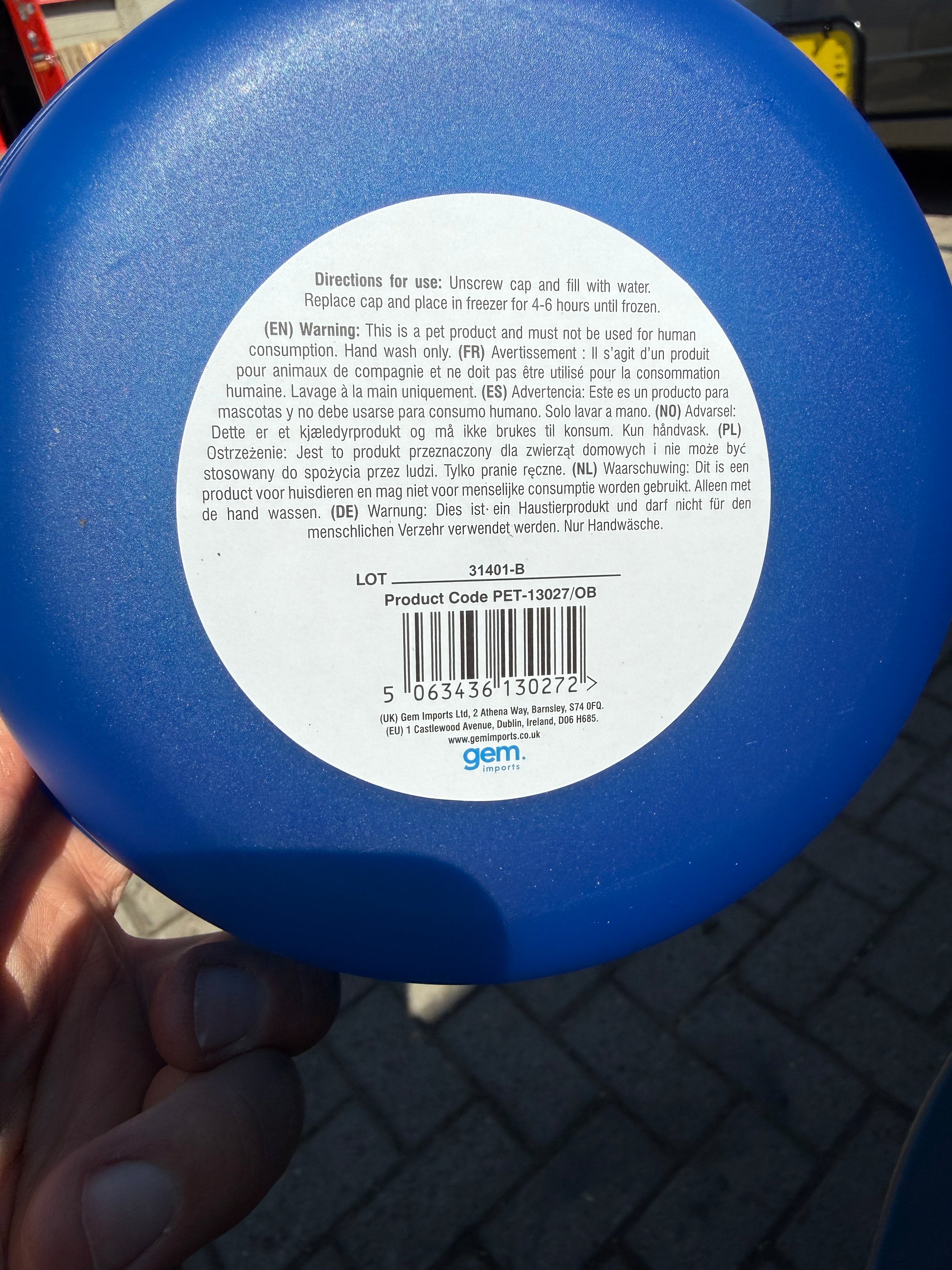 The back of a blue plastic bottle with a white label containing product information, held in a hand against a dark paved background.