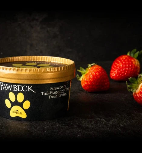 A tub of Pawbeck dog treats on a dark textured surface with fresh strawberries placed behind it. The tub is black with a gold lid and a yellow paw print logo, labeled Strawberry Tail-Waggingly Treat for dogs.